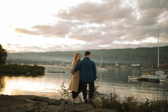 A couple stands by a lakeshore at sunset, with boats and a forested hillside in the background. The woman wears a tan coat and the man a denim jacket. Their pose is calm and intimate, facing the water. by Justin Salem Meyer
