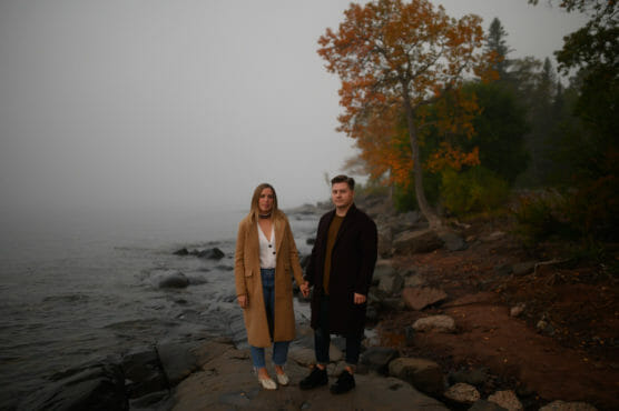 A couple stands on a rocky lakeshore holding hands, with autumn trees in the background and fog over the water. Both wear long coats and look towards the camera. by Justin Salem Meyer