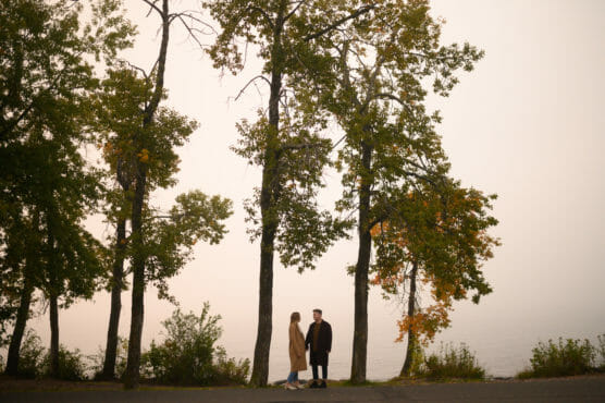 A couple stands facing each other on a path by a lake, surrounded by tall trees with green and yellow leaves on a misty day. by Justin Salem Meyer