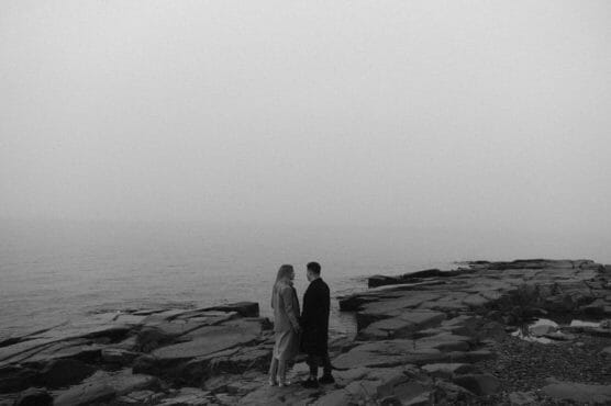 Two people stand facing each other on rocky terrain by the sea, with mist or fog creating a moody, gray atmosphere. The scene appears calm and contemplative. by Justin Salem Meyer