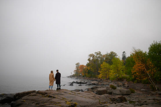 Two people stand on rocky ground near a misty lakeshore, holding hands and facing the water. Trees with autumn foliage line the shoreline under a cloudy, overcast sky. by Justin Salem Meyer