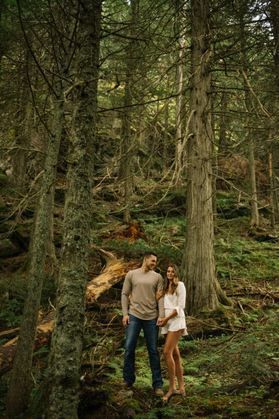 A couple stands close together in a lush, green forest. The man wears jeans and a beige sweater, while the woman wears a white dress. Tall trees and mossy ground surround them in the peaceful woodland setting. by Justin Salem Meyer