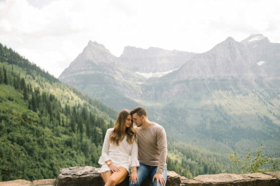 A couple sits close together on a stone ledge, with lush green mountains and a cloudy sky in the background. The woman wears a white dress, and the man wears a beige sweater and jeans. by Justin Salem Meyer