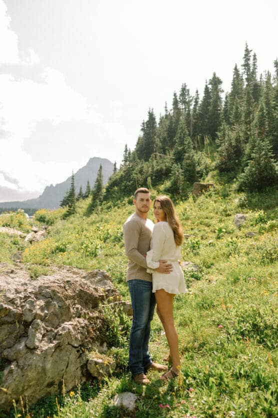A couple stands close together in a sunlit mountain meadow, surrounded by greenery and pine trees, with rocky hills and a distant mountain in the background. by Justin Salem Meyer