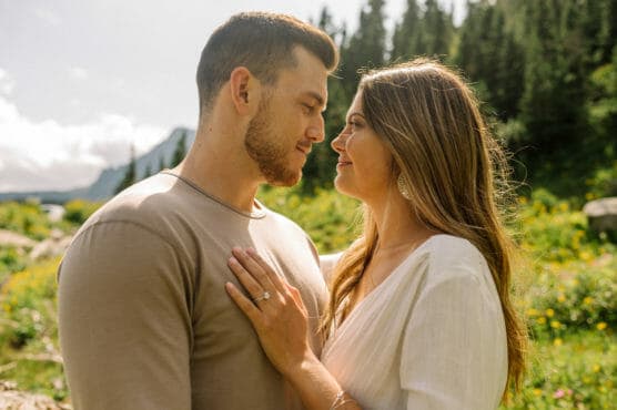 A couple stands close together outdoors, gazing into each others eyes. The woman rests her hand on the mans chest, showing an engagement ring. Sunlight and greenery fill the background, suggesting a scenic, natural setting. by Justin Salem Meyer