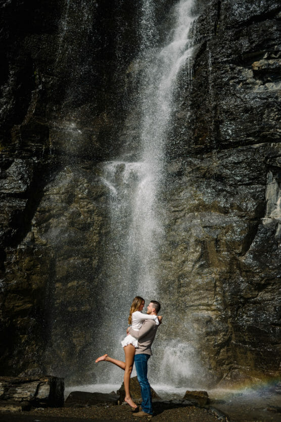 A couple embraces at the base of a tall waterfall, with the man lifting the woman off the ground. Sunlight highlights the falling water and a faint rainbow appears near their feet against the rocky backdrop. by Justin Salem Meyer