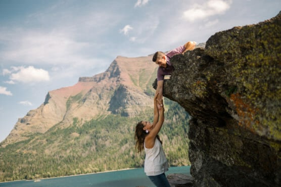 A woman reaches up to help a man who is climbing on a rocky ledge, with scenic mountains and a lake in the background under a partly cloudy sky. by Justin Salem Meyer