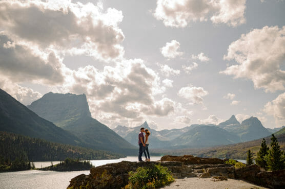 A couple stands close together on a rocky overlook, surrounded by mountains, a lake, and a partly cloudy sky, with sunlight illuminating the scenic landscape. by Justin Salem Meyer