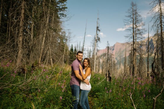 A couple stands close together, smiling in a lush, green field with wildflowers and tall, sparse trees. Mountain peaks and a blue sky with a few clouds are visible in the background. by Justin Salem Meyer