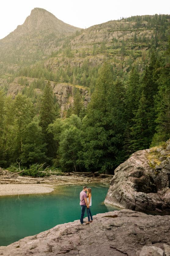 A couple stands on a rocky ledge near a turquoise river, surrounded by dense forest and tall mountains under a hazy sky. by Justin Salem Meyer