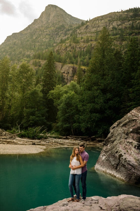 A couple stands on a rocky shore by a turquoise river, surrounded by tall evergreen trees and mountains in the background, embracing each other and smiling peacefully. by Justin Salem Meyer