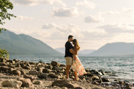A couple stands close together on a rocky lakeshore, surrounded by mountains and water under a sunny, partly cloudy sky. The woman wears a floral dress, and the man is dressed in shorts and a t-shirt. by Justin Salem Meyer