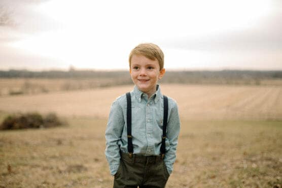 A young boy stands outdoors in a field, wearing a light blue shirt, dark suspenders, and olive green pants. He smiles while looking slightly to the side. The background shows an open landscape under a cloudy sky. by Justin Salem Meyer