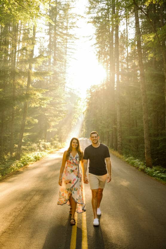 A couple walks hand in hand down a sunlit road surrounded by tall trees, with sunlight streaming through the forest and casting a warm glow. by Justin Salem Meyer
