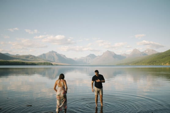 A woman in a floral dress and a man in dark clothes stand in the shallow water of a calm lake, surrounded by mountains under a clear sky with scattered clouds. by Justin Salem Meyer
