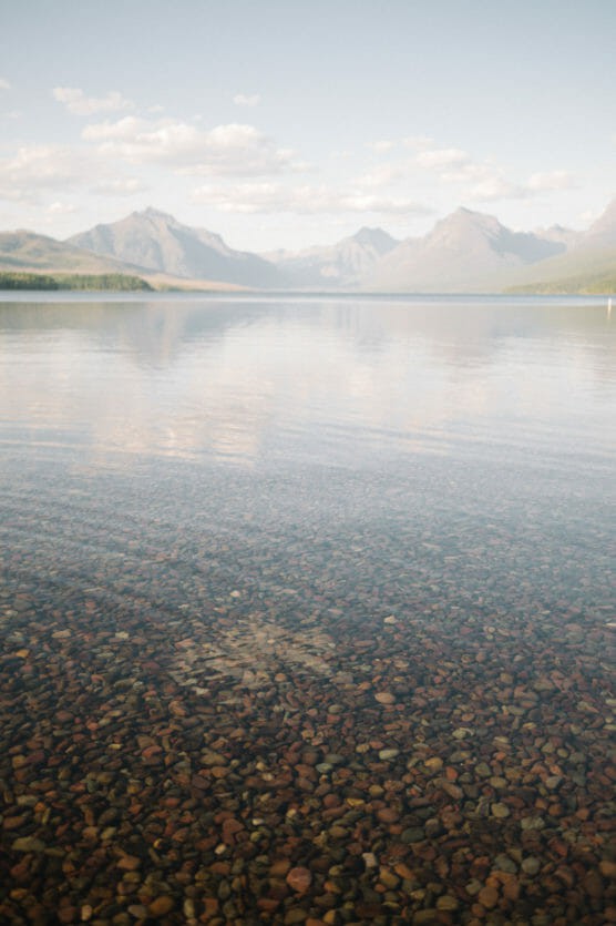 Clear, shallow lake with multicolored pebbles visible underwater in the foreground, surrounded by distant mountains under a blue sky with scattered clouds. by Justin Salem Meyer