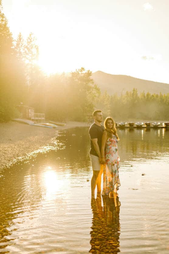 A couple stands together in shallow lake water at sunset, surrounded by trees and distant mountains. The woman wears a floral dress, and the man wears shorts and a t-shirt. Warm sunlight creates a golden, serene atmosphere. by Justin Salem Meyer