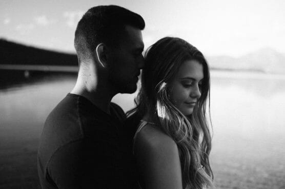 A black and white photo of a man standing behind a woman by a calm lake. The man faces the woman, while she looks down and smiles softly. Mountains and sky appear blurred in the background. by Justin Salem Meyer