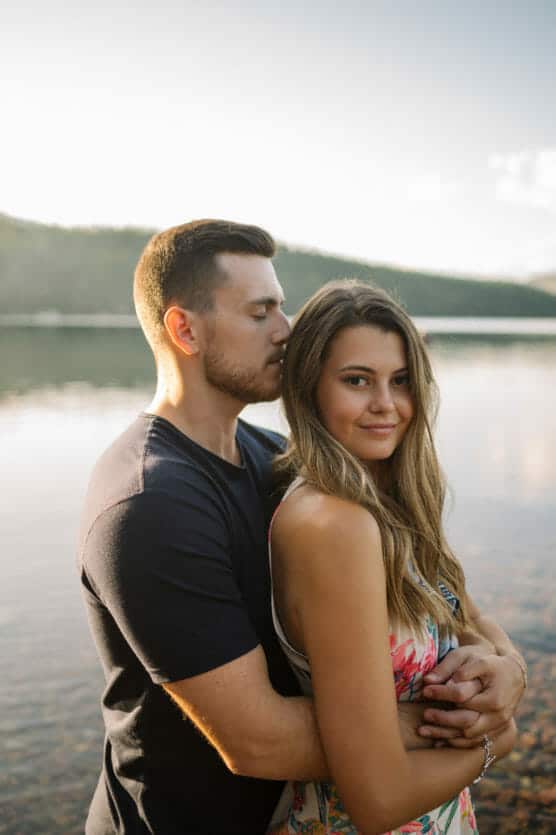 A man embraces a woman from behind as they stand by a calm lake. The woman smiles softly at the camera, while the man rests his face near her head. Trees and hills are visible in the background under soft sunlight. by Justin Salem Meyer
