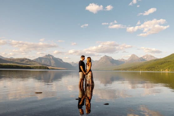 A couple stands holding hands on a calm lake with mountains and a blue sky with scattered clouds in the background, their reflections visible in the water. by Justin Salem Meyer