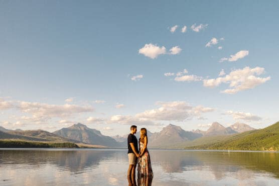 A couple stands holding hands in shallow water with mountains and a clear blue sky with scattered clouds in the background. The scene is tranquil and sunlit, reflecting the landscape in the water. by Justin Salem Meyer
