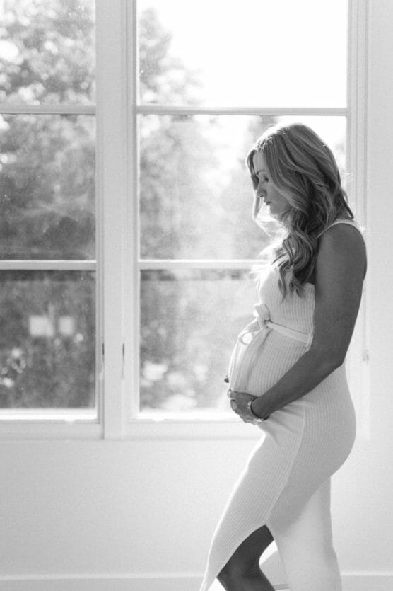 A pregnant woman in a fitted dress stands in profile by a large sunlit window, gently cradling her belly and smiling softly. The scene is in black and white. by Justin Salem Meyer