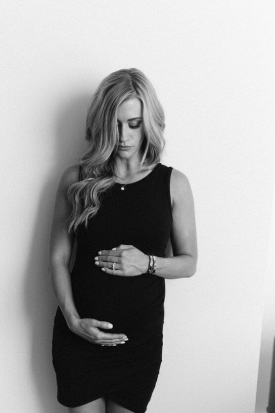A pregnant woman in a sleeveless black dress stands against a white wall, looking down and gently cradling her baby bump with both hands. The image is in black and white. by Justin Salem Meyer