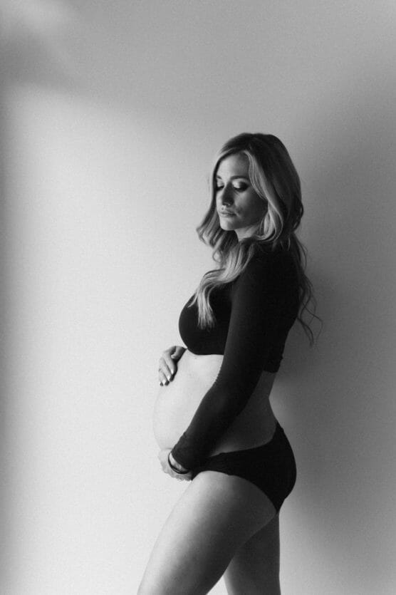A black and white photo of a pregnant woman standing against a plain wall, wearing a long-sleeve crop top and underwear, with one hand on her belly and looking down thoughtfully. by Justin Salem Meyer