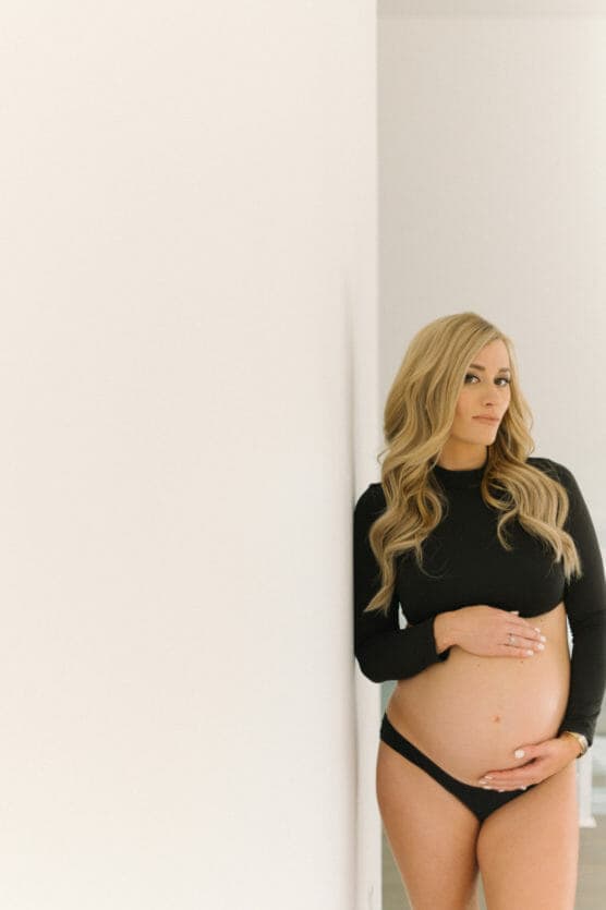 A pregnant woman with long blonde hair, wearing a black long-sleeve top and black underwear, stands indoors leaning against a white wall, gently holding her belly and looking at the camera. by Justin Salem Meyer