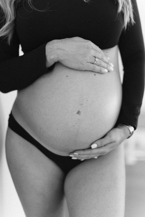 A black and white photo shows a pregnant person in a black top and underwear, gently cradling their bare belly with both hands. Their face is not visible. by Justin Salem Meyer