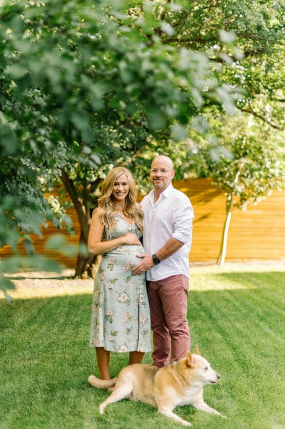 A smiling couple stands together in a sunny green yard, the woman in a floral dress holding her pregnant belly. A tan and white dog lies on the grass in front of them, and leafy trees fill the background. by Justin Salem Meyer