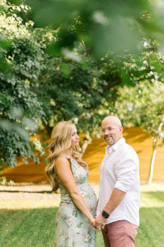 A pregnant woman in a floral dress holds hands with a man in a white shirt and pink pants. They stand outdoors on grass, with trees and a wooden fence in the background, looking at each other. by Justin Salem Meyer