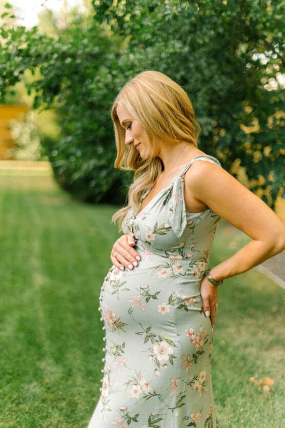 A pregnant woman in a floral dress stands outdoors on green grass, holding her baby bump and looking down with a gentle smile. Lush green trees form the background. by Justin Salem Meyer