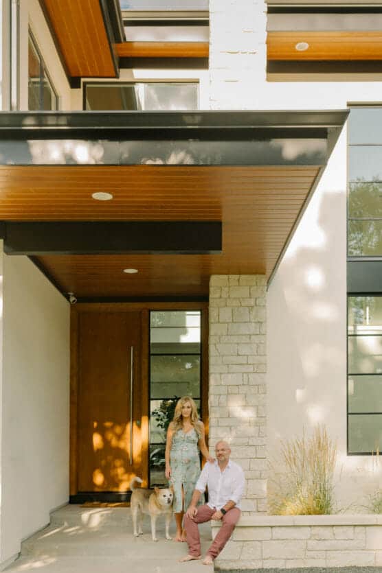 A woman and a man with a dog pose outside a modern house with large windows, a wooden door, and stone and wood accents. Sunlight creates soft shadows on the homes façade. by Justin Salem Meyer