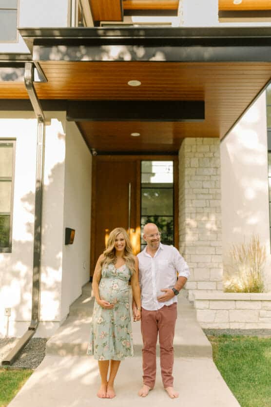 A pregnant woman in a floral dress and a man in a white shirt and pants stand barefoot, holding hands and smiling outside the entrance of a modern house. by Justin Salem Meyer