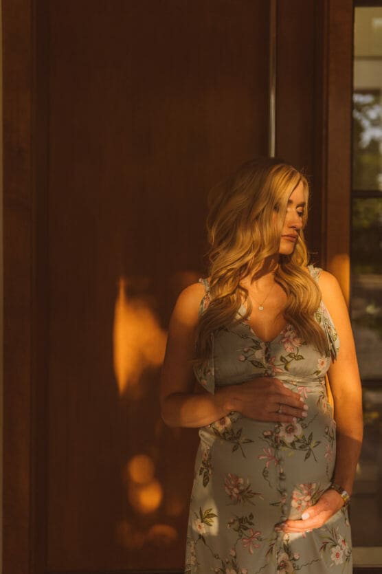 A pregnant woman with long blonde hair stands in warm sunlight, wearing a light floral dress and gently cradling her belly with one hand, looking down with a serene expression. by Justin Salem Meyer