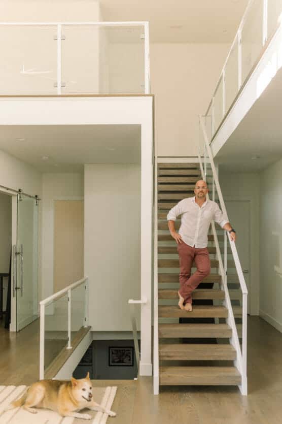 A man in a white shirt and pink pants stands barefoot on a wooden staircase inside a modern, bright home with glass railings. A dog lies on a rug at the bottom of the stairs. by Justin Salem Meyer