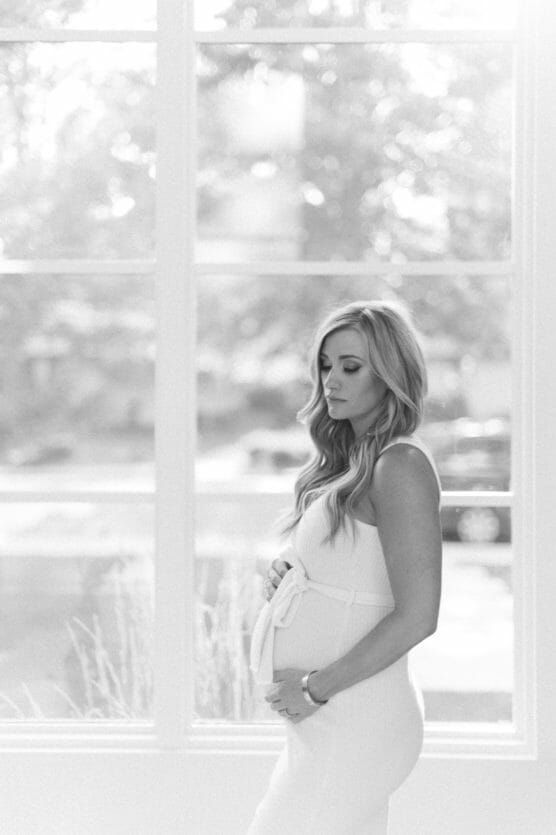 A black and white photo of a pregnant woman in a white dress, standing sideways by a large window, gently holding her belly and looking down with a serene expression. Sunlight softly illuminates the scene. by Justin Salem Meyer