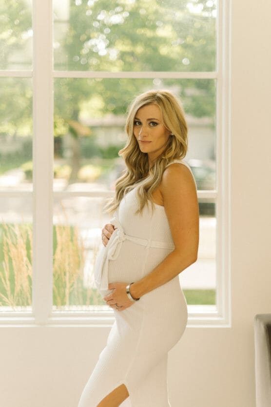 A pregnant woman in a white sleeveless dress stands indoors by a large window, gently cradling her belly and looking at the camera, with soft natural light and greenery visible outside. by Justin Salem Meyer