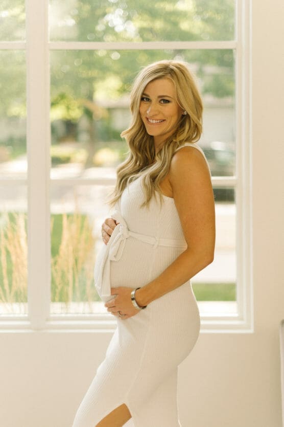 A smiling pregnant woman in a white dress stands indoors by a sunlit window, gently cradling her baby bump with one hand. Greenery and sunlight are visible outside. by Justin Salem Meyer