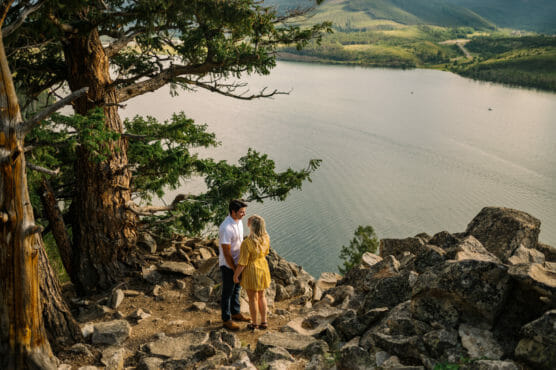 A couple stands on rocky terrain beneath a large tree, overlooking a calm lake and green hills in the distance on a sunny day. by Justin Salem Meyer