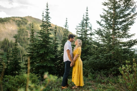 A couple stands close together, holding hands and facing each other in a forested mountain area. The woman wears a yellow dress, and the man wears a white shirt and dark pants. Tall evergreen trees and hills surround them. by Justin Salem Meyer