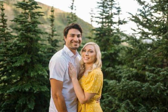 A couple stands smiling together outdoors in front of green pine trees; the man wears a short-sleeve white shirt and the woman wears a yellow dress, with sunlight filtering through the trees behind them. by Justin Salem Meyer