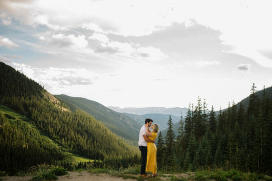 A couple stands on a mountain overlook, embracing each other. The woman wears a yellow dress, and the landscape features lush green pine forests and distant mountain peaks under a partly cloudy sky. by Justin Salem Meyer