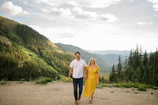 A man in a white shirt and jeans holds hands with a woman in a yellow dress as they walk outdoors with green mountains, trees, and a partly cloudy sky in the background. by Justin Salem Meyer