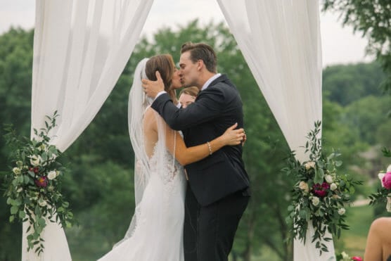 A bride and groom kiss under a decorated white arch during an outdoor wedding ceremony, surrounded by greenery and floral arrangements, with a person standing behind them. by Justin Salem Meyer