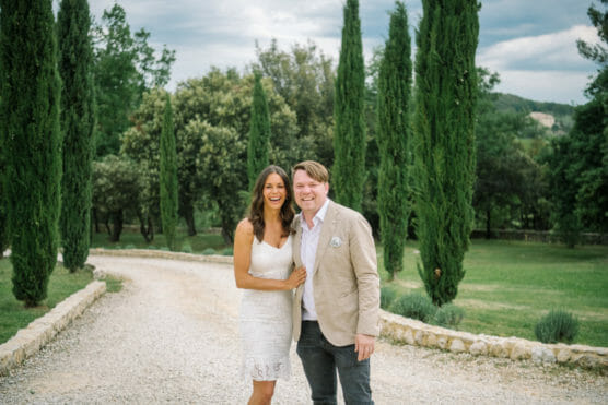 A smiling couple stands on a gravel path lined with tall cypress trees and greenery. The woman wears a white dress and the man wears a beige jacket and blue shirt. They are outside on a cloudy day. by Justin Salem Meyer