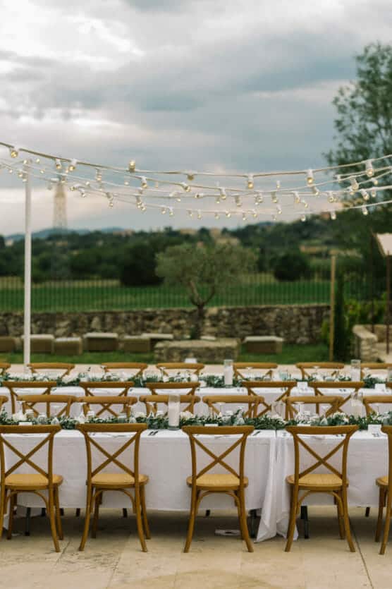 A long outdoor dining table with wooden chairs is set with white tablecloths and greenery. String lights hang above, and the table overlooks a grassy field and stone wall under a cloudy sky. by Justin Salem Meyer