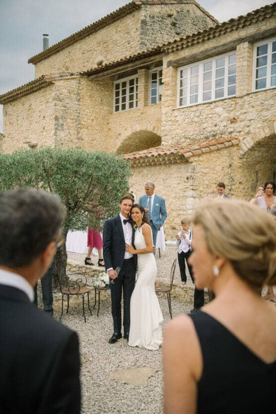 A bride in a white dress and a groom in a tuxedo stand together, smiling and posing for a photo outside a rustic stone building, surrounded by wedding guests in formal attire. by Justin Salem Meyer