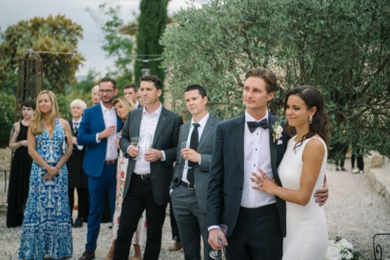 A bride in a white dress and groom in a tuxedo stand arm in arm, smiling, surrounded by well-dressed guests outdoors in a garden setting, some holding drinks and watching the couple attentively. by Justin Salem Meyer